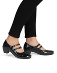 Black patent mary jane shoe with two gold buckle straps and a low block heel worn with black pants on model.