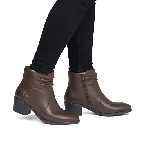 A pair of women's brown leather ankle boots with a ruched shaft, low block heel, and side zipper, worn by a model wearing black pants.