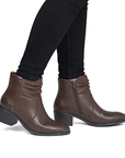 A pair of women's brown leather ankle boots with a ruched shaft, low block heel, and side zipper, worn by a model wearing black pants.