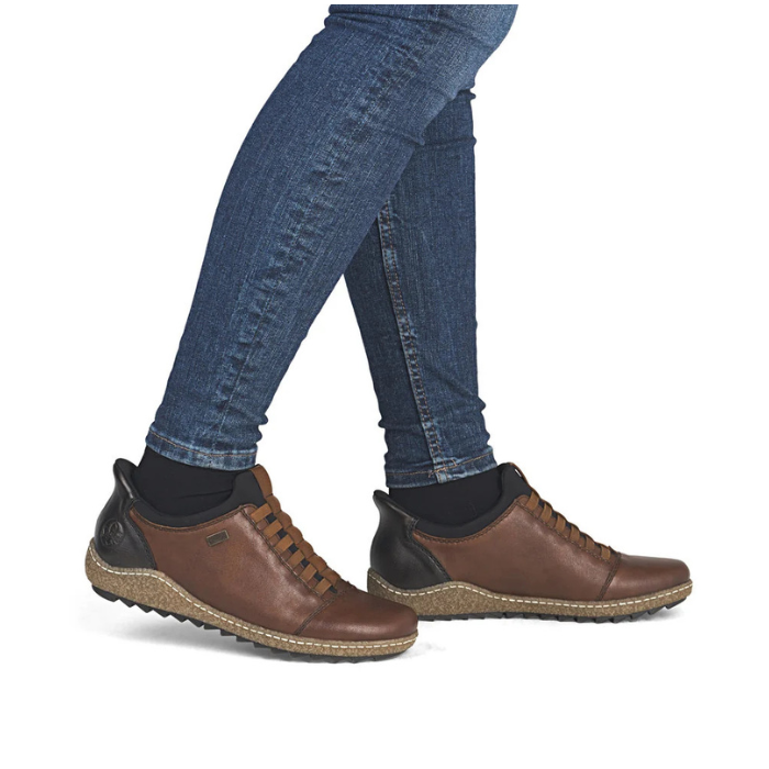 A person wearing blue skinny jeans and brown leather lace-up shoes with black heel accents, shown against a plain white background.