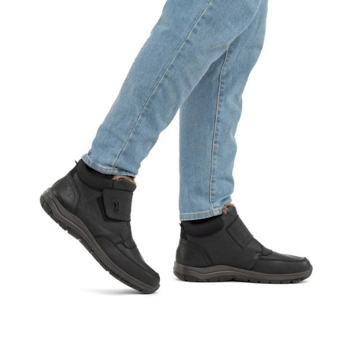 Black ankle boot with Velcro strap and blue jeans worn by man.