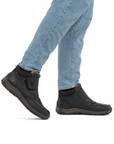 Black ankle boot with Velcro strap and blue jeans worn by man.