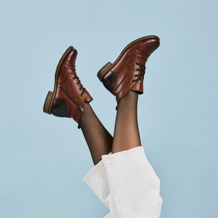 Brown leather shoes worn with black tights and a white sock against a light blue background.
