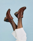 Brown leather shoes worn with black tights and a white sock against a light blue background.