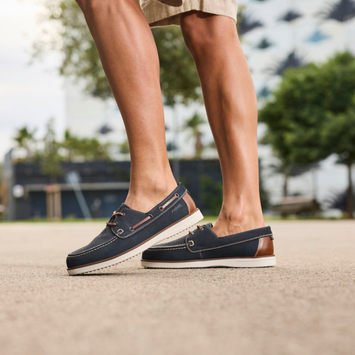 Navy blue boat shoes with brown accents worn by a person on a blurred outdoor background.