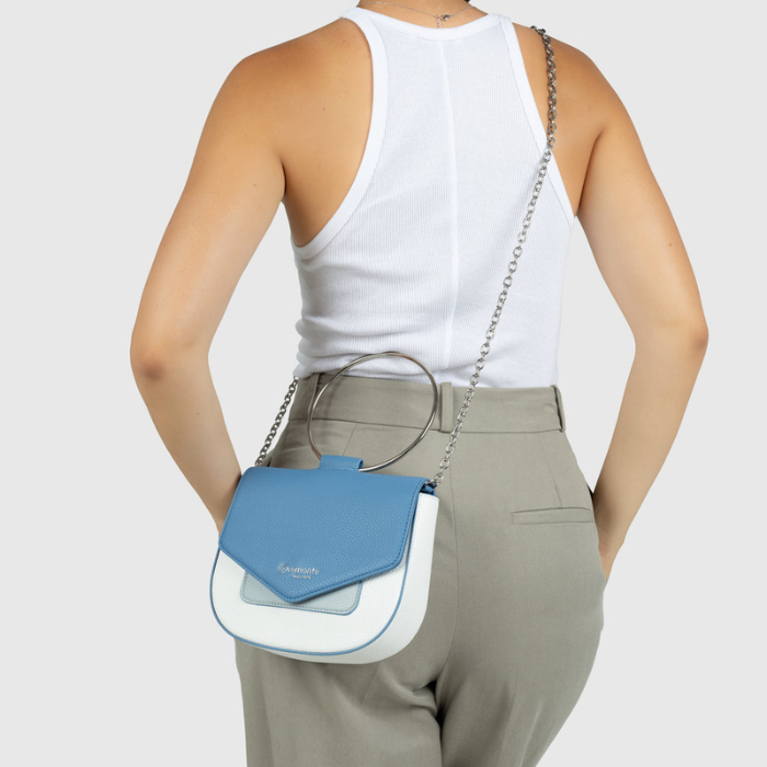 A woman shown from the back wearing a white tank top and light gray trousers, carrying a small crossbody bag with a blue flap, white body, and silver chain strap.