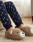 Child wearing navy pajama pants with white stars and brown bear slippers on a carpeted floor.