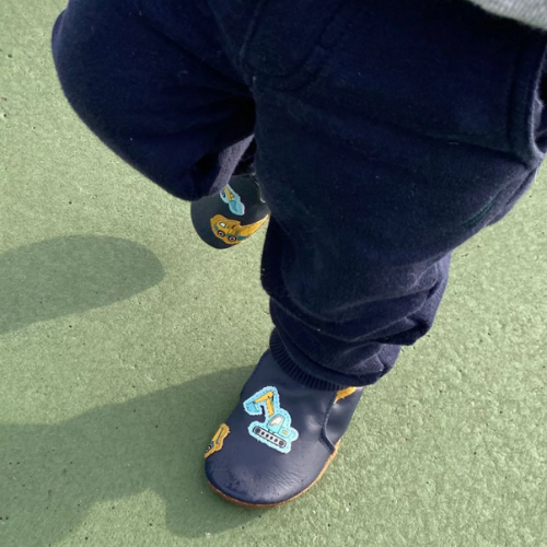 Child's feet wearing navy blue baby shoes with construction vehicle pattern standing on green floor.