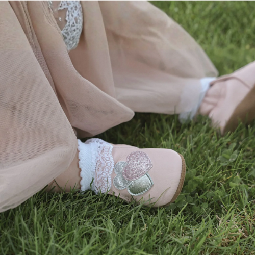 Child's feet peaking out of a pink dress wearing white socks and pink leather shoes with sparkly hearts on vamps.