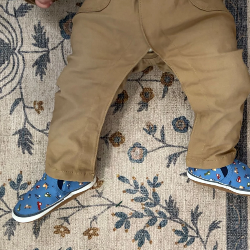 Baby wearing beige pants and blue canvas shoes with vehicle pattern on a patterned rug.