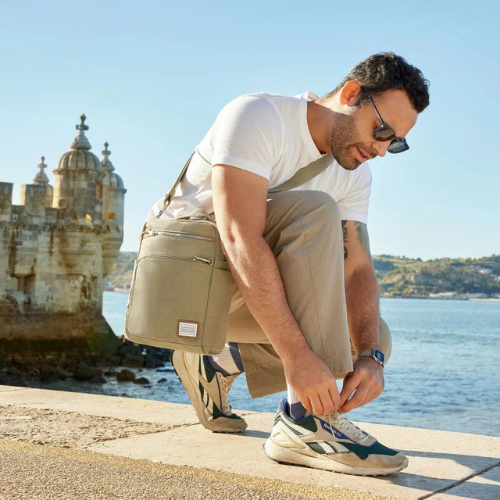 Man sitting by a waterfront with a beige bag, wearing sunglasses and a white shirt.