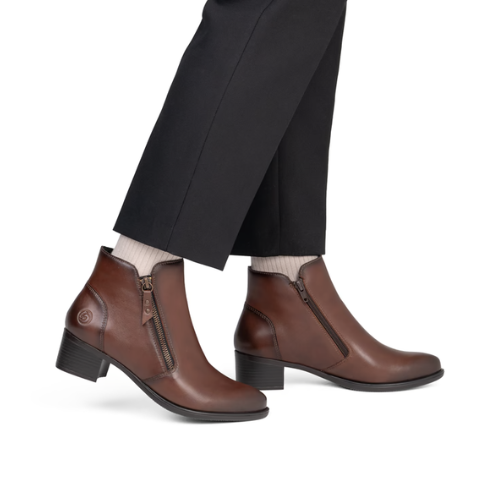 A pair of brown leather ankle boots with side zippers, shown on a model wearing black trousers and white socks.