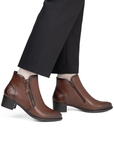 A pair of brown leather ankle boots with side zippers, shown on a model wearing black trousers and white socks.