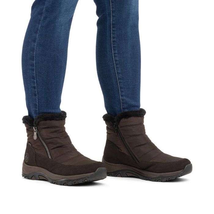 A pair of dark brown ankle winter boots with black faux-fur trim and side zippers, shown on a model wearing blue jeans.