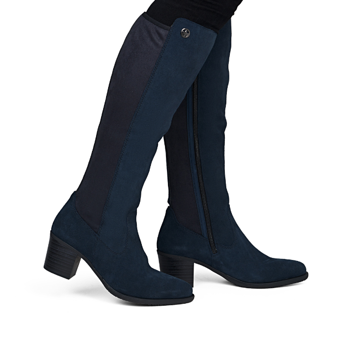 A pair of knee-high boot in dark navy blue suede, with a low block heel worn by woman in black jeans
