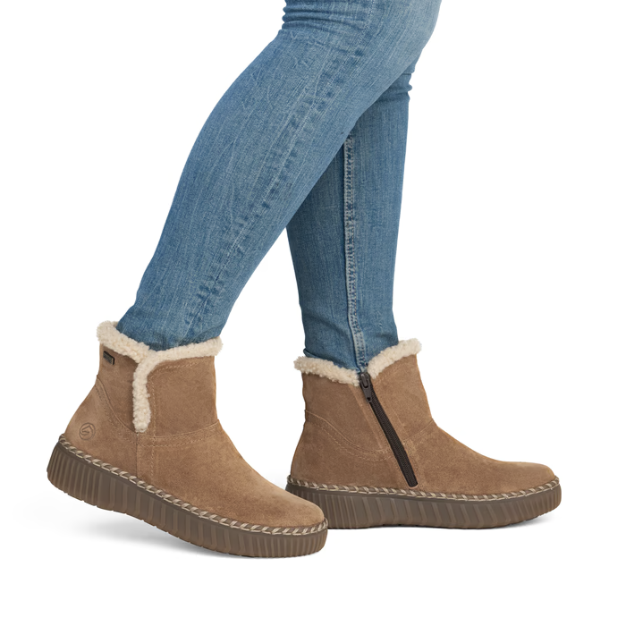 A person wearing tan suede ankle boots with white shearling lining and blue jeans, shown from the knees down.