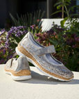 Pair of silver Mary-Jane shoes on a surface with flowers in the background.