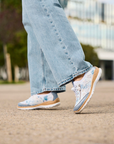 Person wearing white and light blue sneakers with jeans outdoors.