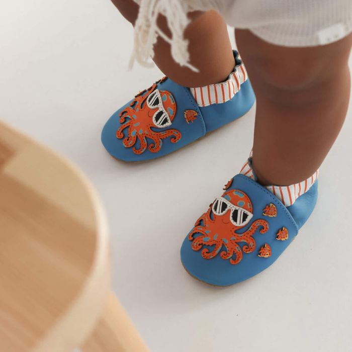 Baby wearing blue leather shoes with orange octopus and fish on them.