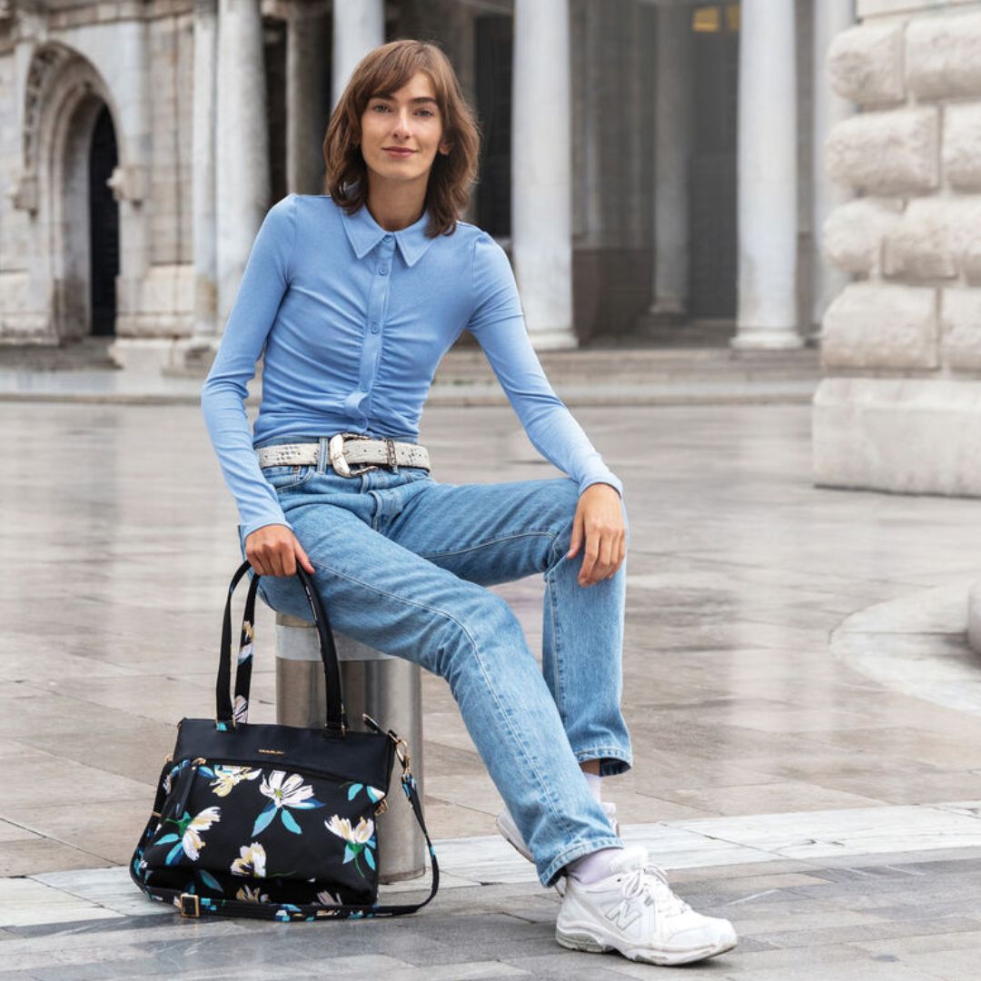 Women in a blue top and jeans is holding a black with floral pattern tote bag.