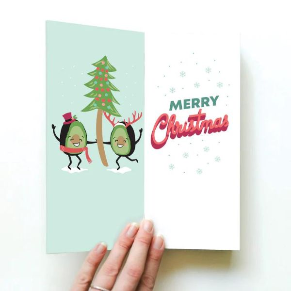 Card with two avocado people holding a Christmas Tree. Text reads Merry Christmas.