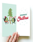 Card with two avocado people holding a Christmas Tree. Text reads Merry Christmas.