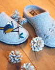 Songbird Slippers on a wooden floor near some pinecones.