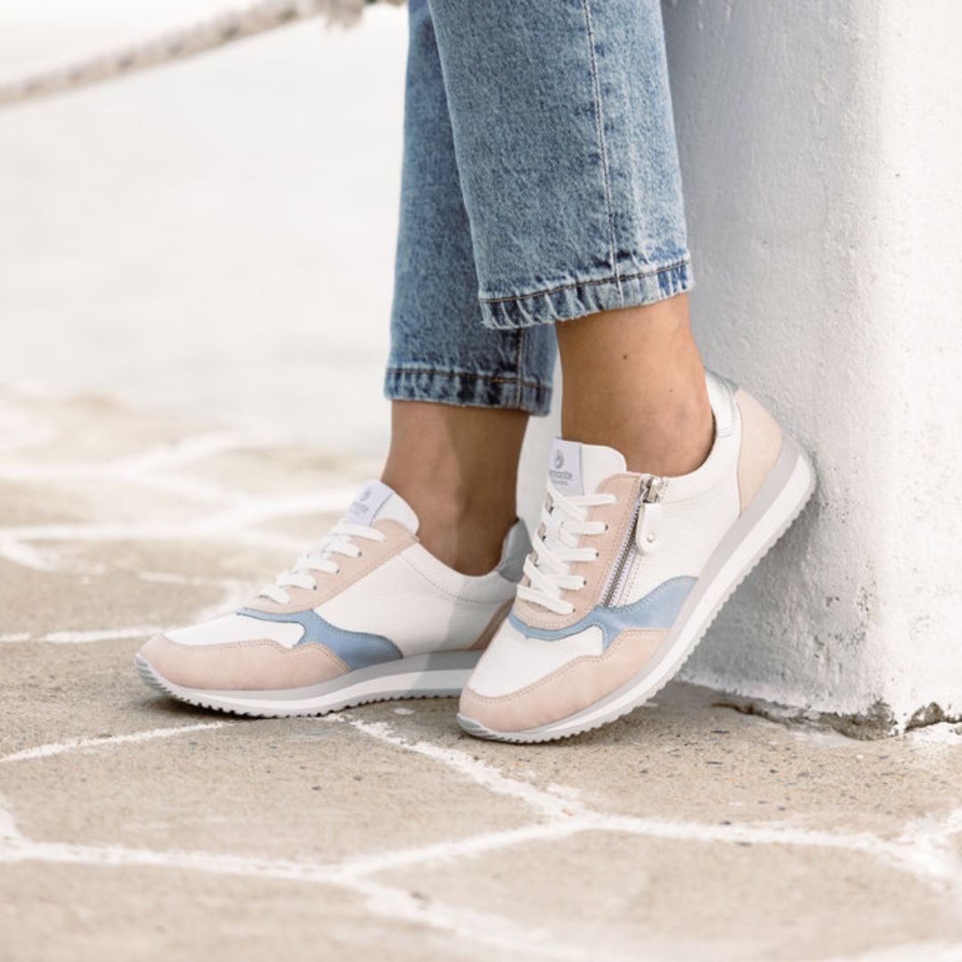 Women in jeans wearing white, blue and pink sneaker with grey and white outsole. Sneaker has white laces and zipper closure.
