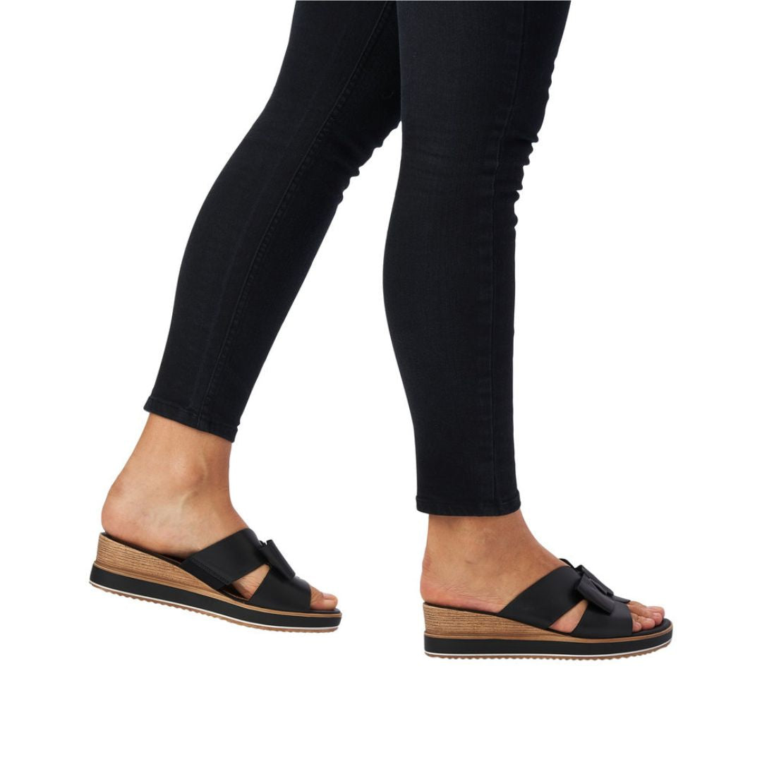 Legs in black jeans wearing black leather slide wedge sandal with bow.