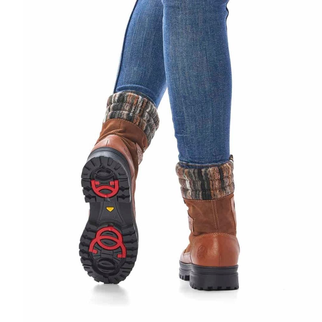 Women in jeans wearing brown lace up boot with yarn knit cuff, inside zipper and black outsole with red collapsible ice cleats..