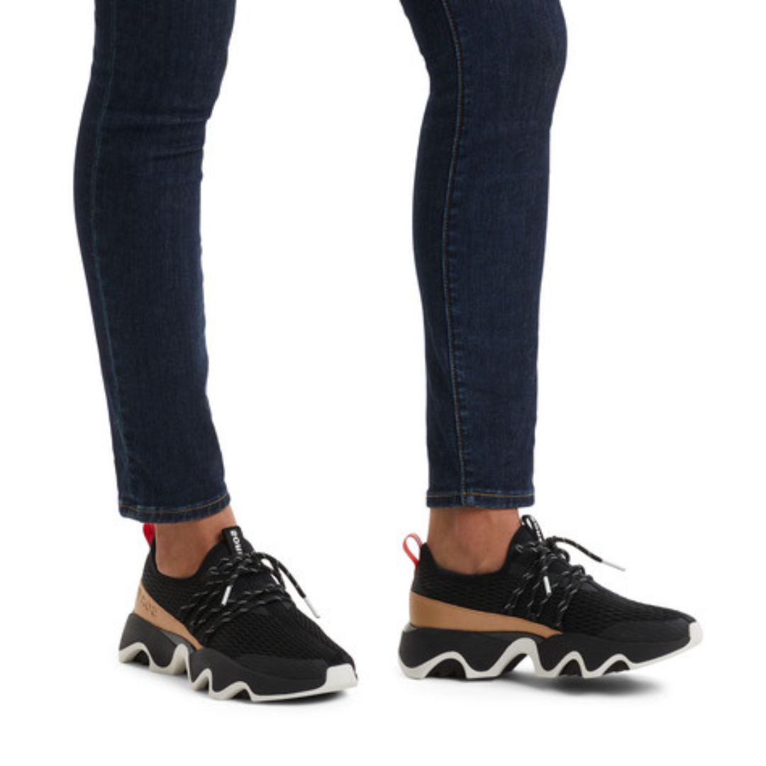 Legs in dark jeans wearing black mesh sneakers with brown accent and thick black and white outsoles.