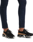 Legs in dark jeans wearing black mesh sneakers with brown accent and thick black and white outsoles.