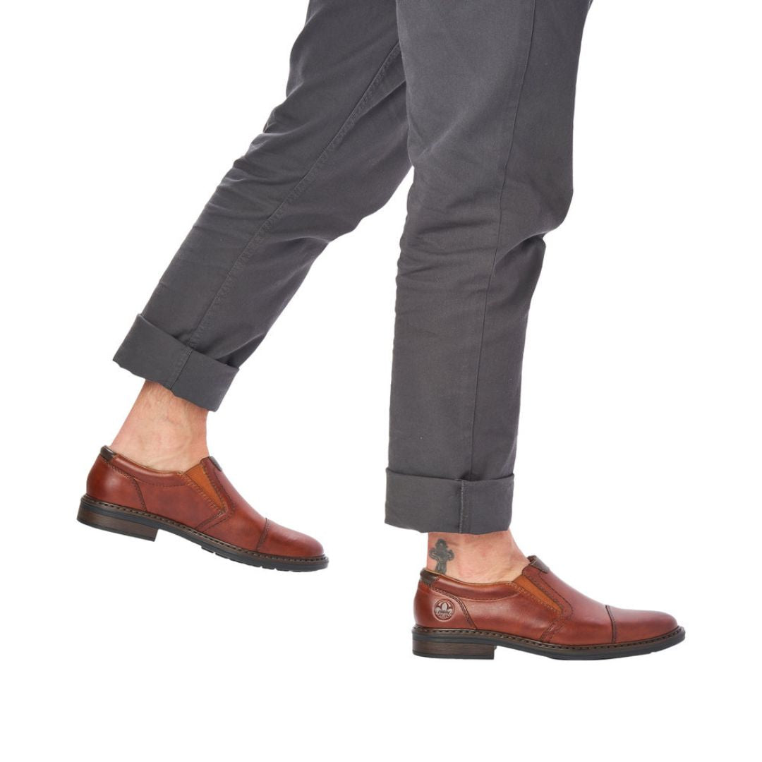 Man in grey pants wearing brown slip on dress shoes with Rieker logo on heel.